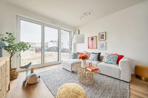 Blick ins eingerichtete Wohnzimmer des Musterhauses. Durch die Balkontür hindurch ist die Baustelle im Hintergrund zu sehen.